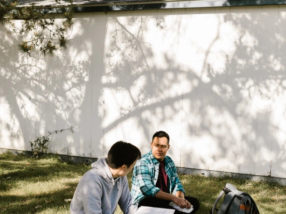 two people sitting in the grass beneath a tree with books and backpacks on the ground