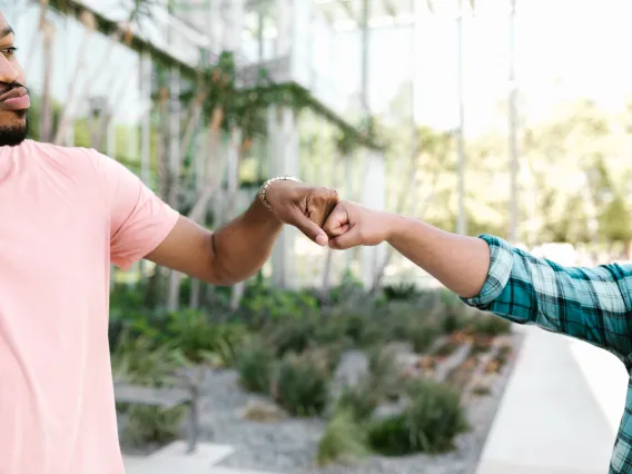 two people bumping fists outdoors with a glass building and trees in the background