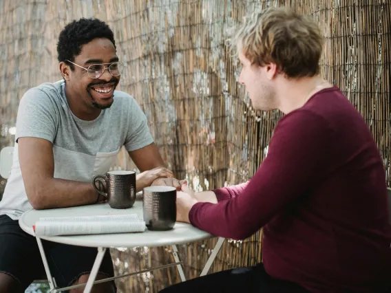 two people sitting at a table talking with mugs in front of them