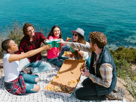 group of friends raising cups into the air and eating pizza sitting outdoors by the water