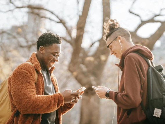 two people standing outdoors looking at cell phones and smiling together