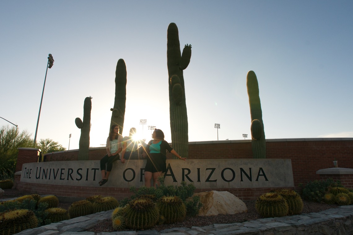 group of students setting in front of a University of Arizona sign with the sun setting behind them