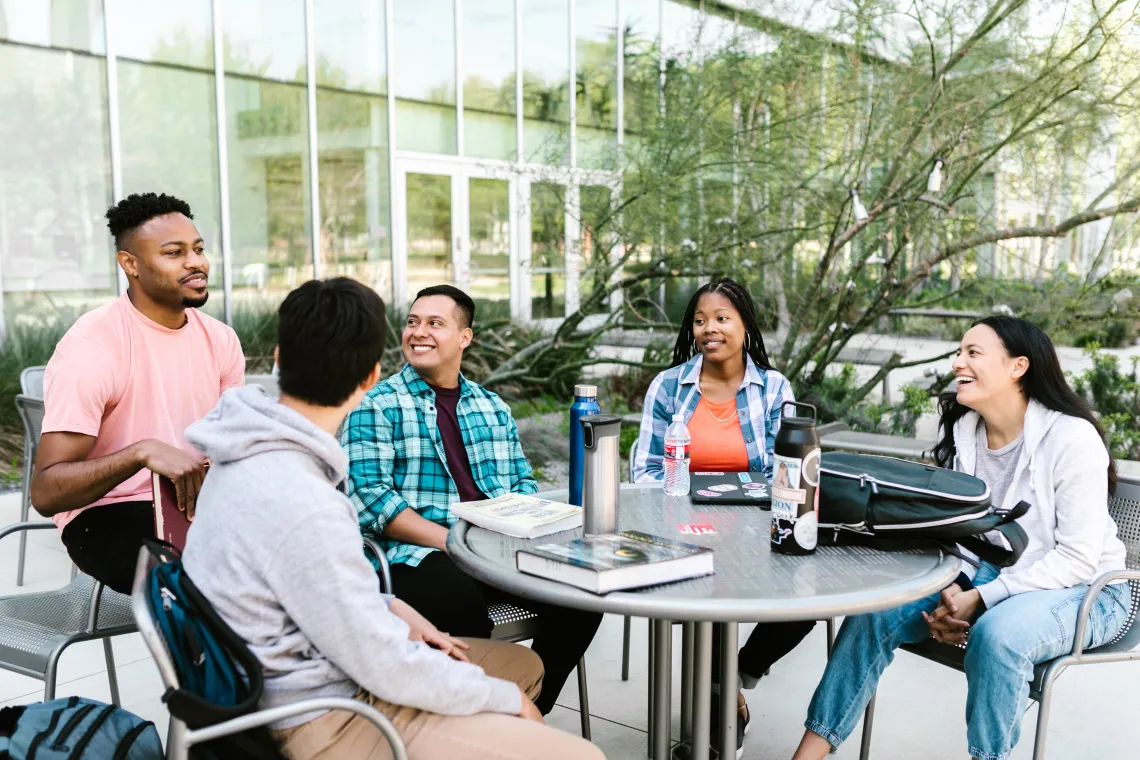 group of students sitting at an outdoor table with books and binders on the table and a building in the background