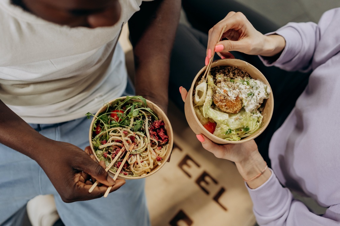 overhead view of two people sitting on a wooden box eating bowls of vegetables and noodles
