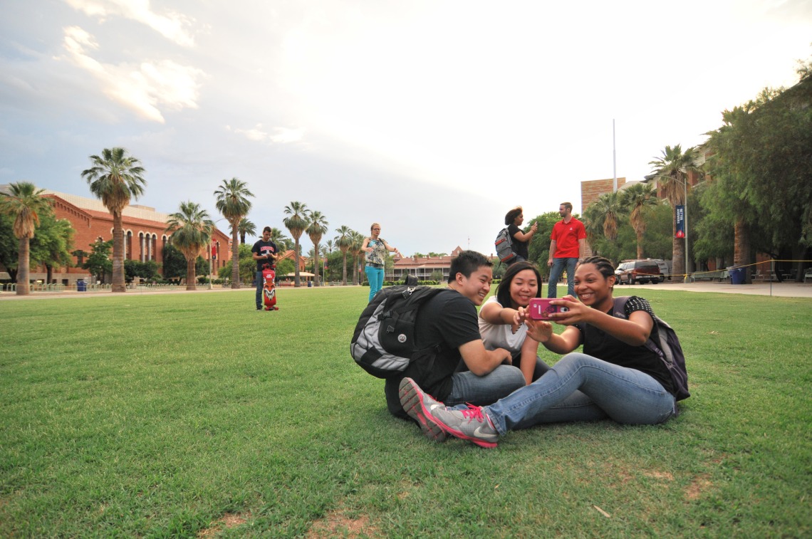 group of students sitting on the lawn and looking at a phone on the University of Arizona campus