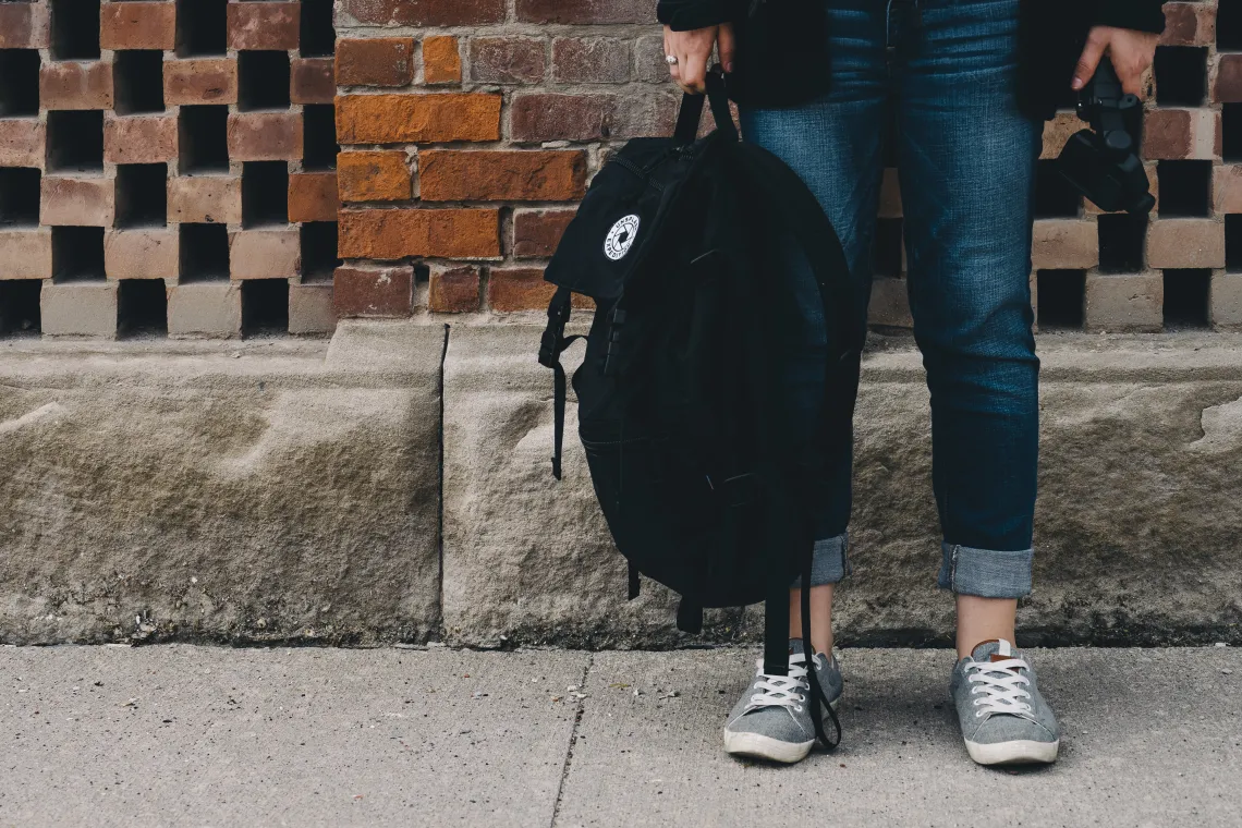 person standing on a sidewalk in front of a brick wall and holding a backpack to the side